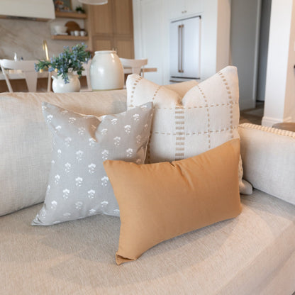 Three decorative pillows in neutral tones arranged on a beige sofa: one with white floral pattern, one with tan geometric gri