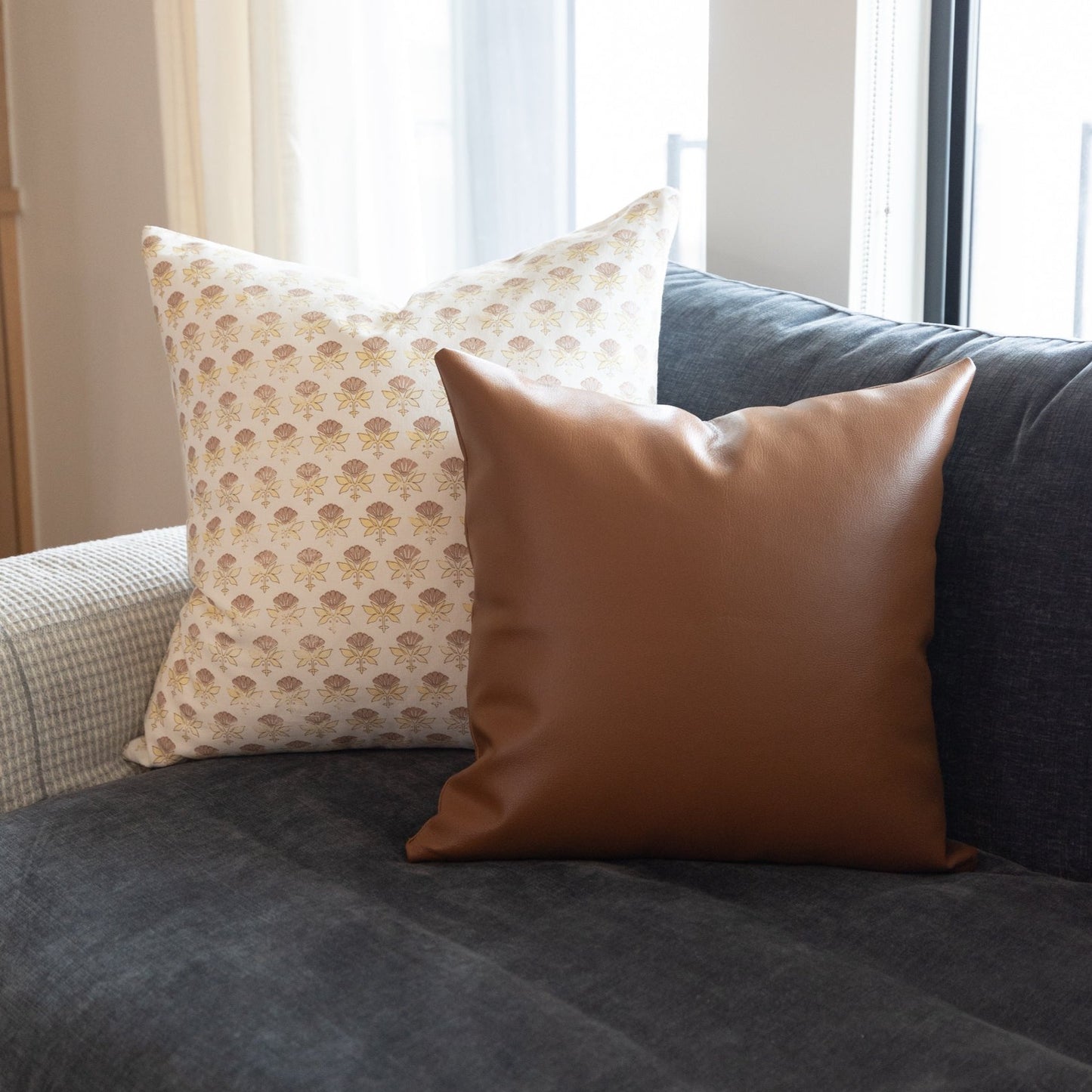 White and tan floral patterned pillow displayed on a charcoal sofa with brown leather pillow beside it.