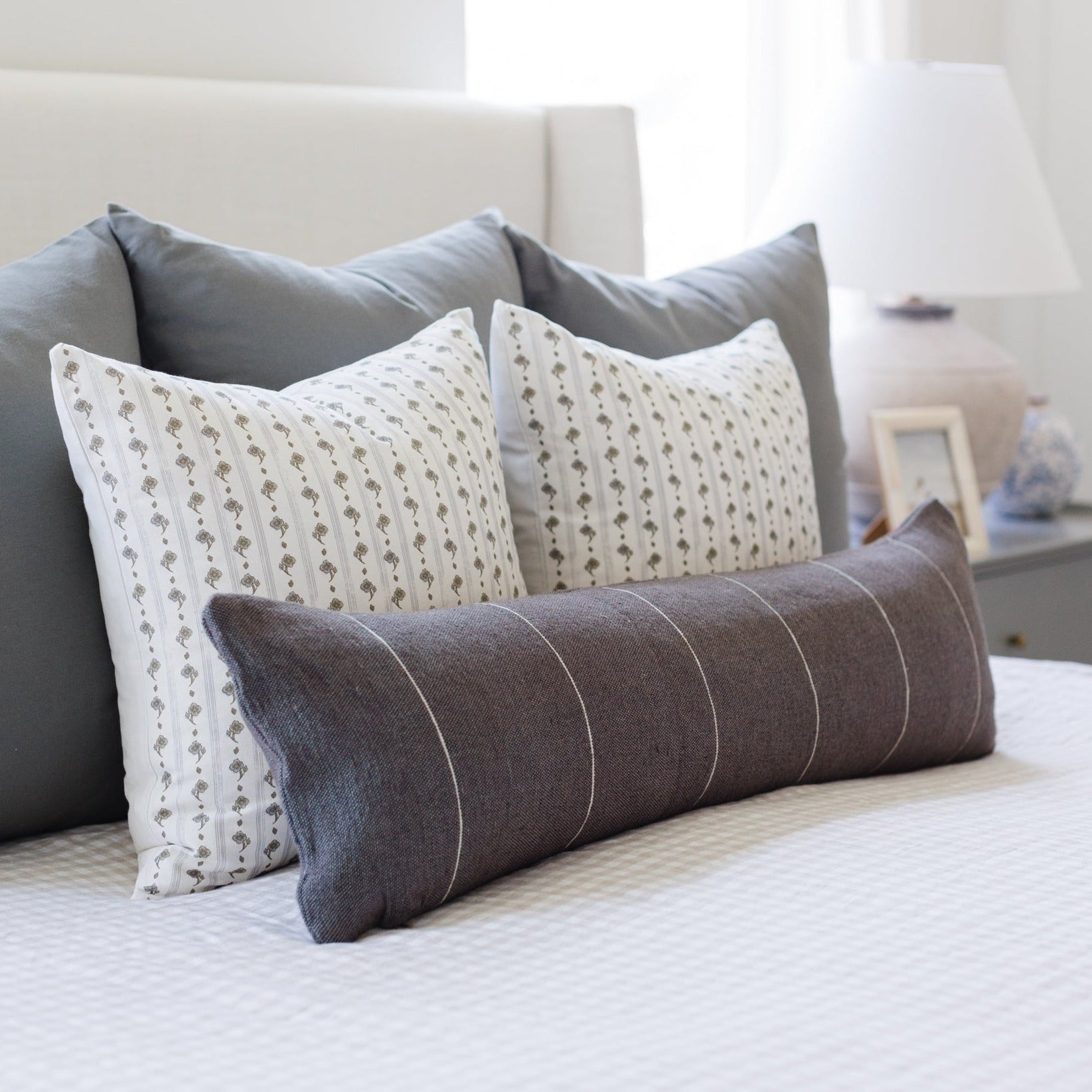 Three decorative pillows on a bed: two cream with small floral pattern and one charcoal with vertical stripe design.