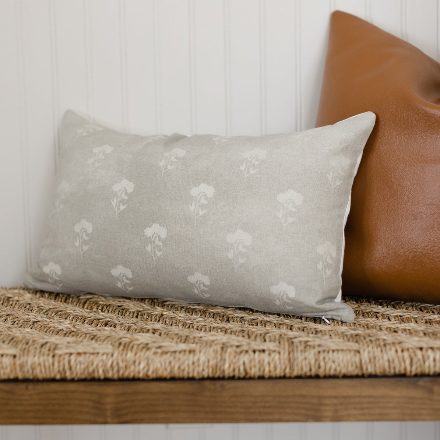 Gray lumbar pillow with white floral pattern, displayed on a woven bench next to a brown leather pillow.