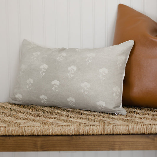 Gray lumbar pillow with white floral pattern, displayed on a woven bench next to a brown leather pillow.