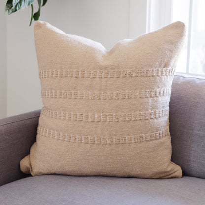 Tan woven pillow with horizontal textured stripe pattern, displayed on gray upholstered sofa.
