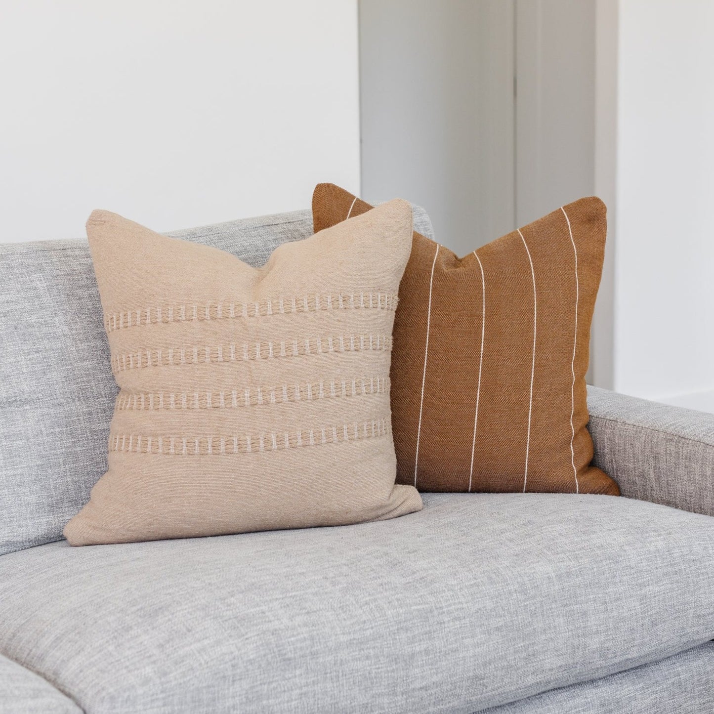 Tan throw pillow with geometric stripe and dot pattern details, displayed on a gray upholstered sofa.