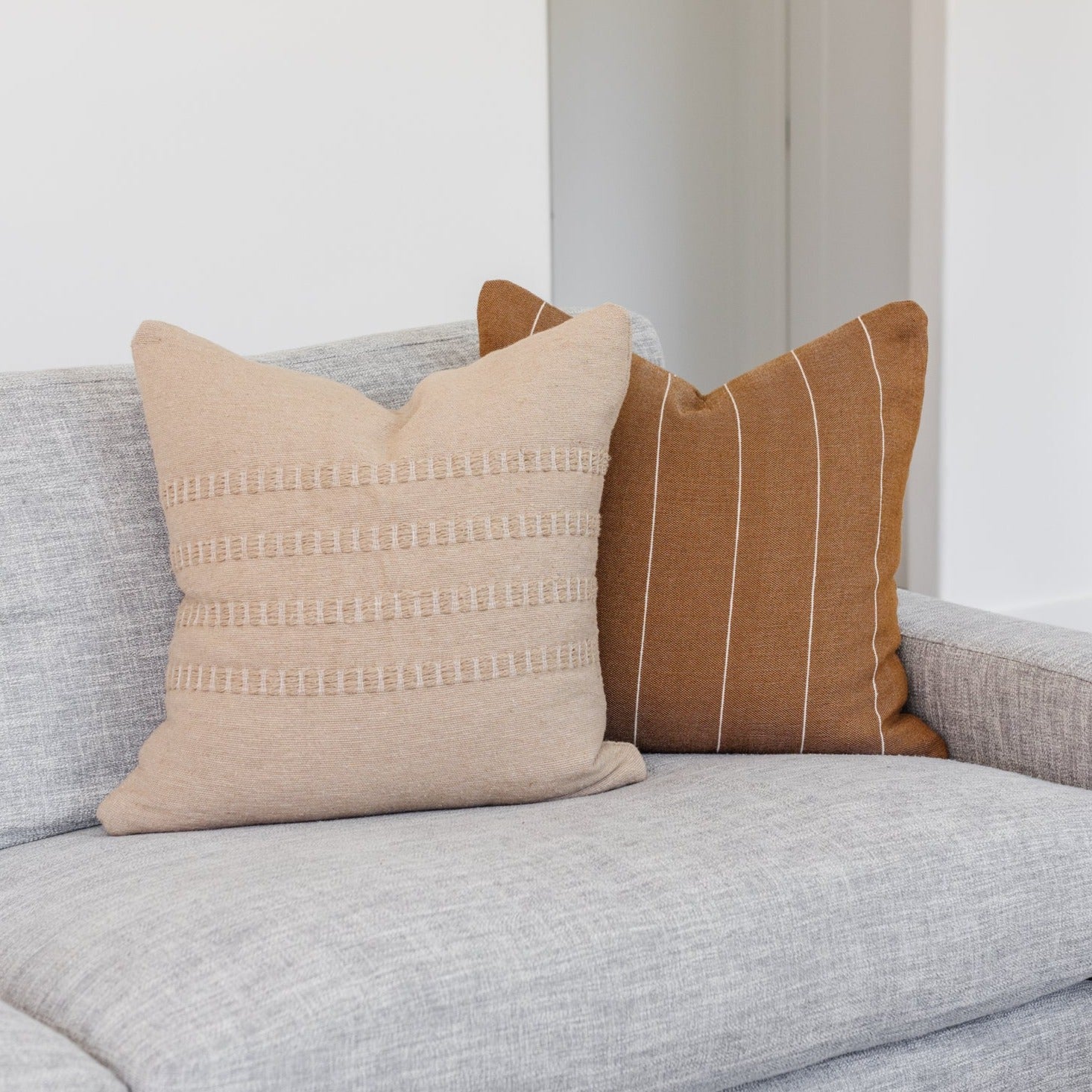 Tan throw pillow with geometric stripe and dot pattern details, displayed on a gray upholstered sofa.