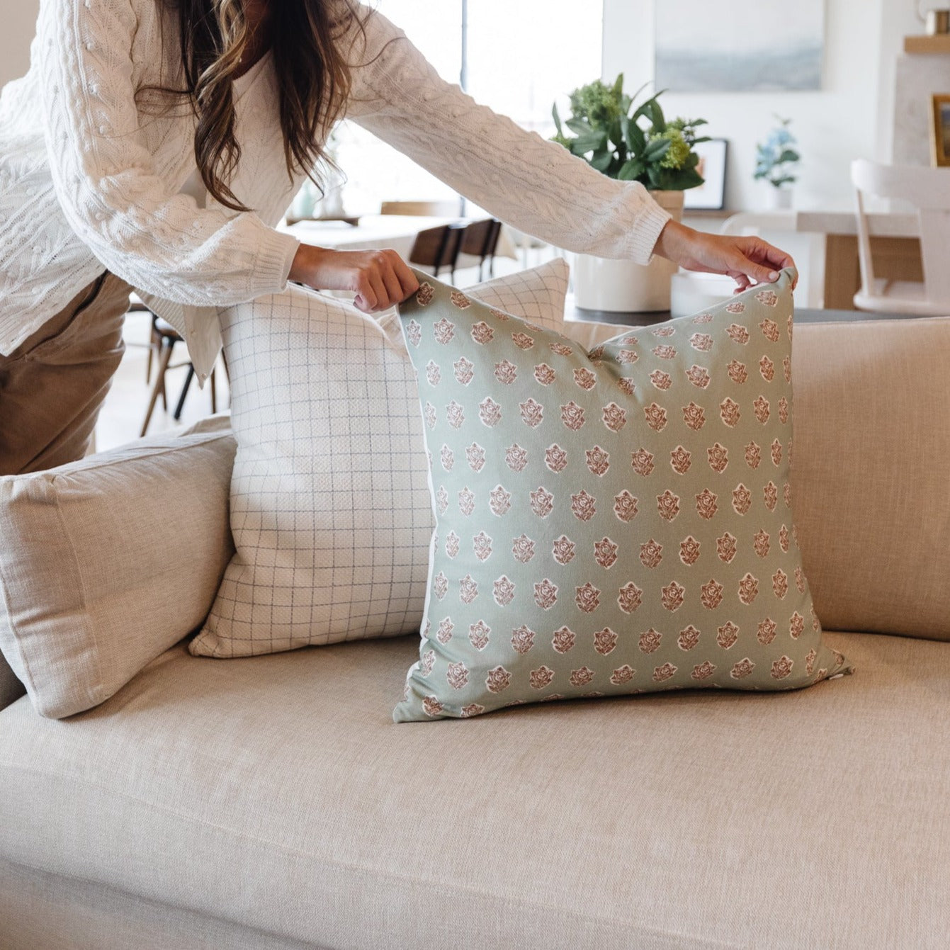 Four decorative pillows in various neutral and patterned fabrics arranged on a beige sofa with a woman displaying one.