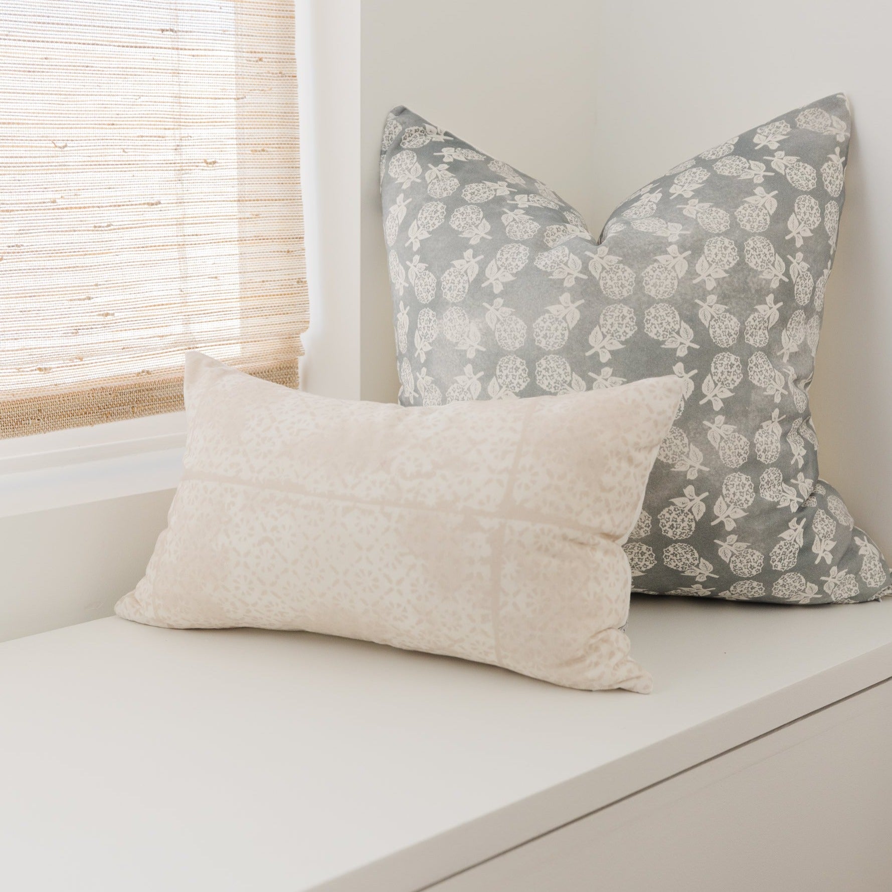 Gray pillow with cream floral print and cream solid rectangular pillow on white shelf.