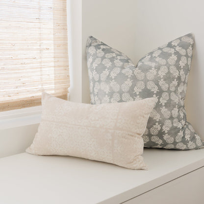 Cream lumbar pillow and gray pillow with white floral pattern displayed on a white surface near a window.