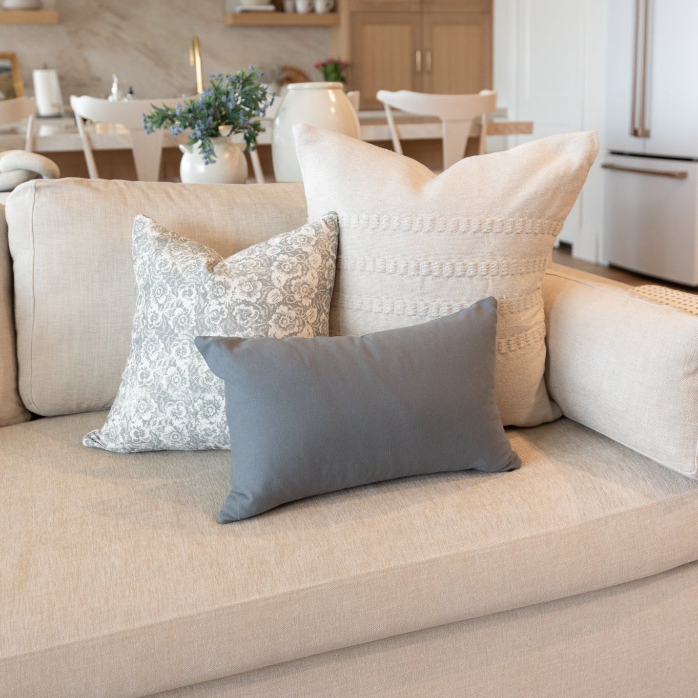 Three decorative pillows on a beige sofa: gray rectangle, white textured square, and gray patterned square.