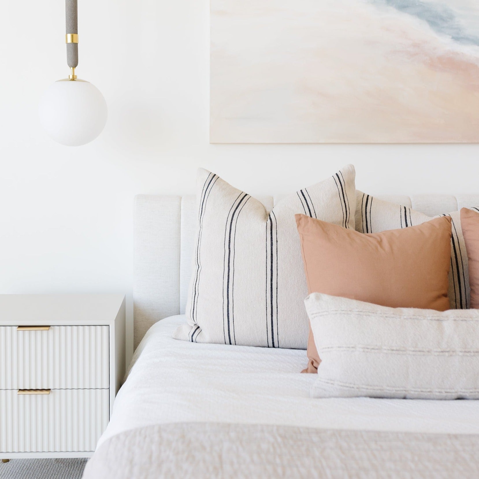 Bedroom styled with white bedding, striped and terracotta pillows, pendant light, and nightstand.