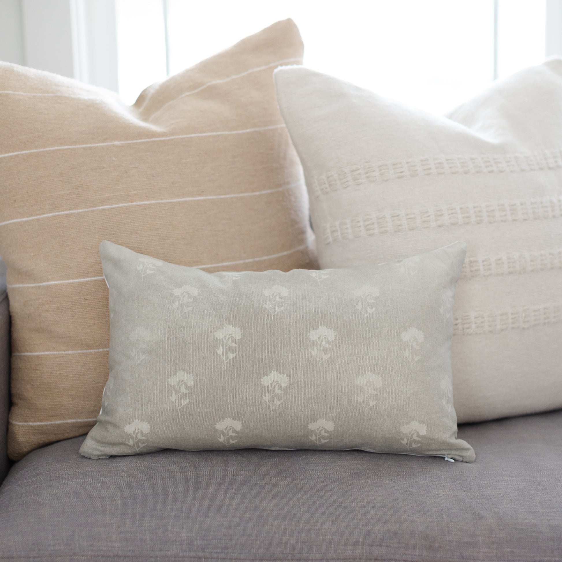 Gray lumbar pillow with cream floral pattern, displayed among coordinating neutral pillows on a sofa.