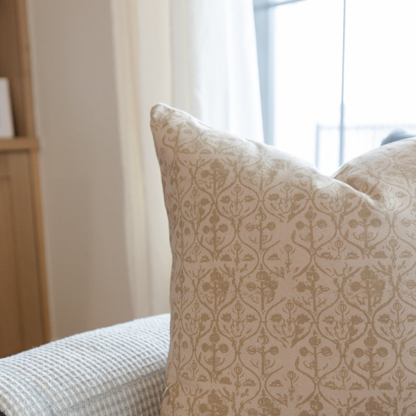 Beige pillow cover with repeating damask floral pattern in tan tones, displayed on a bed near a window.