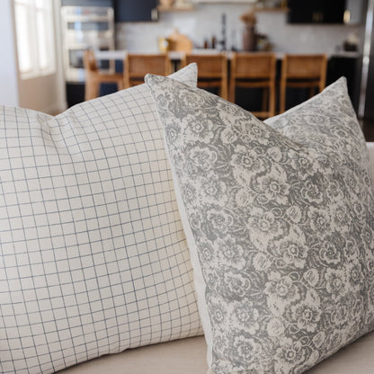 Two gray and cream patterned throw pillows, one with floral damask print and one with grid pattern, displayed on a sofa.