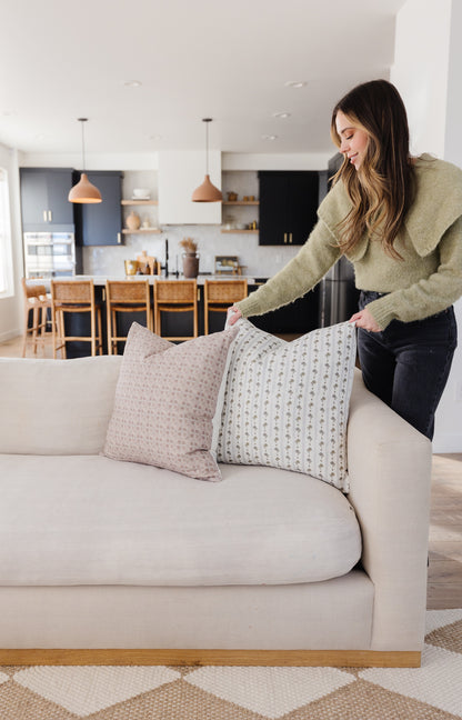 Woman arranging two patterned throw pillows on a cream-colored sofa in a modern kitchen setting.