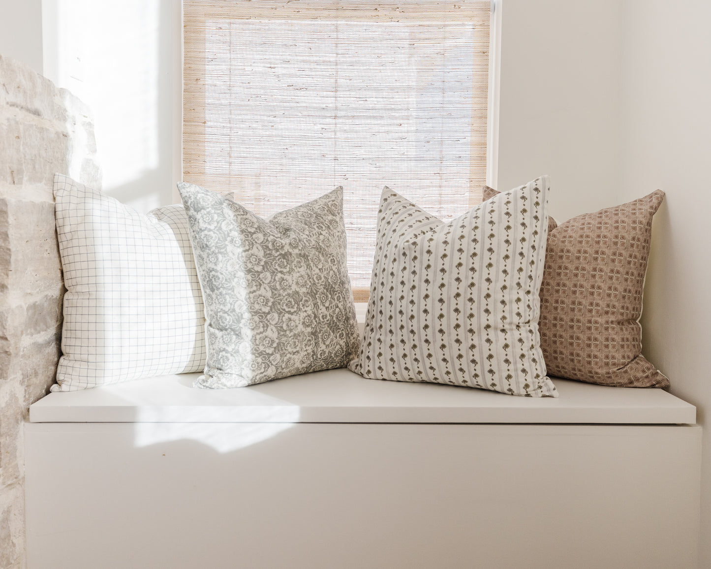 Four patterned throw pillows in neutral tones with grid, floral, striped, and geometric designs arranged on a white window se