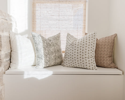 Four patterned throw pillows in neutral tones with grid, floral, striped, and geometric designs arranged on a white window se