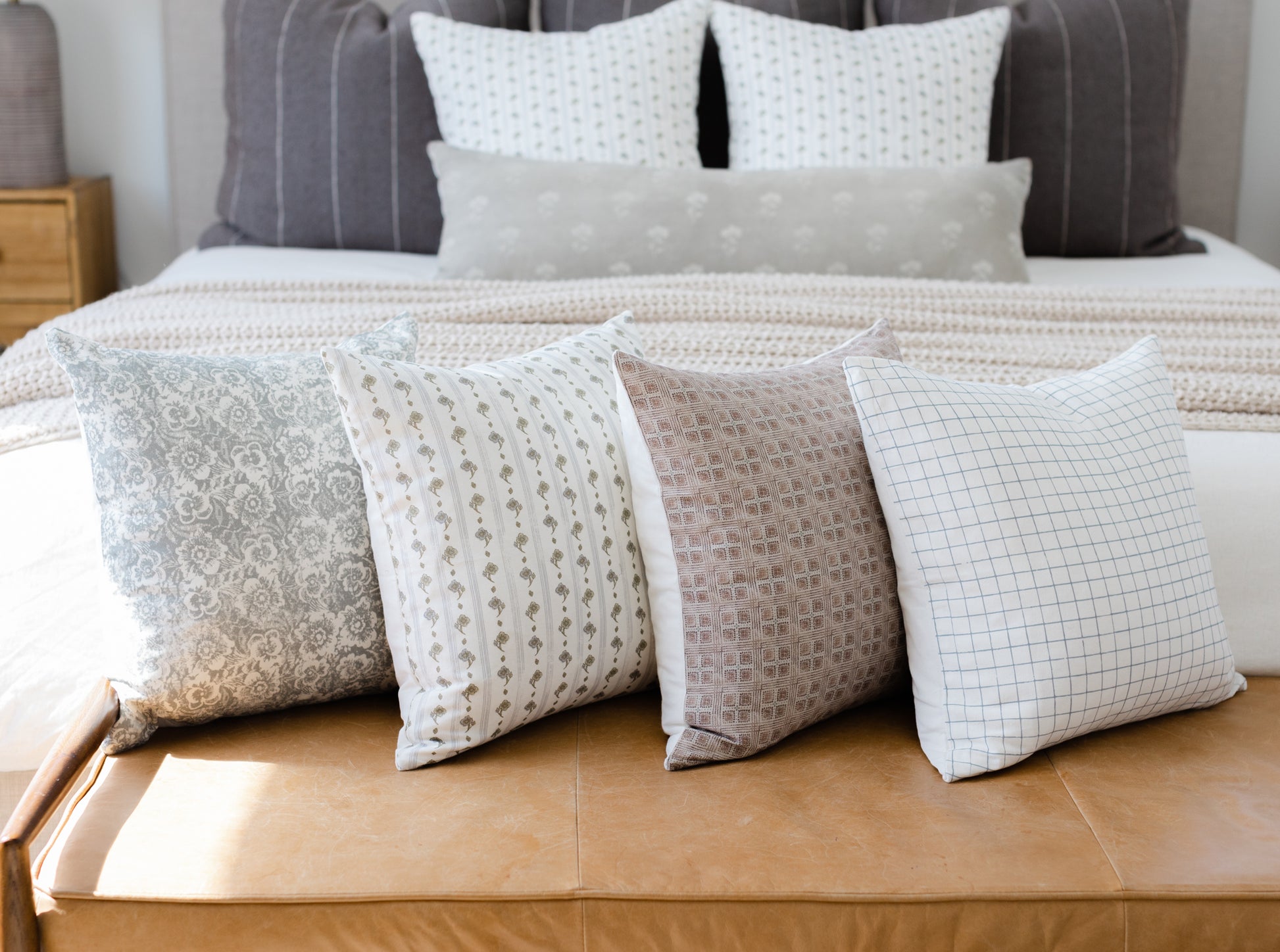 Four decorative pillows in neutral tones with floral, dotted, textured, and grid patterns displayed on a bed.