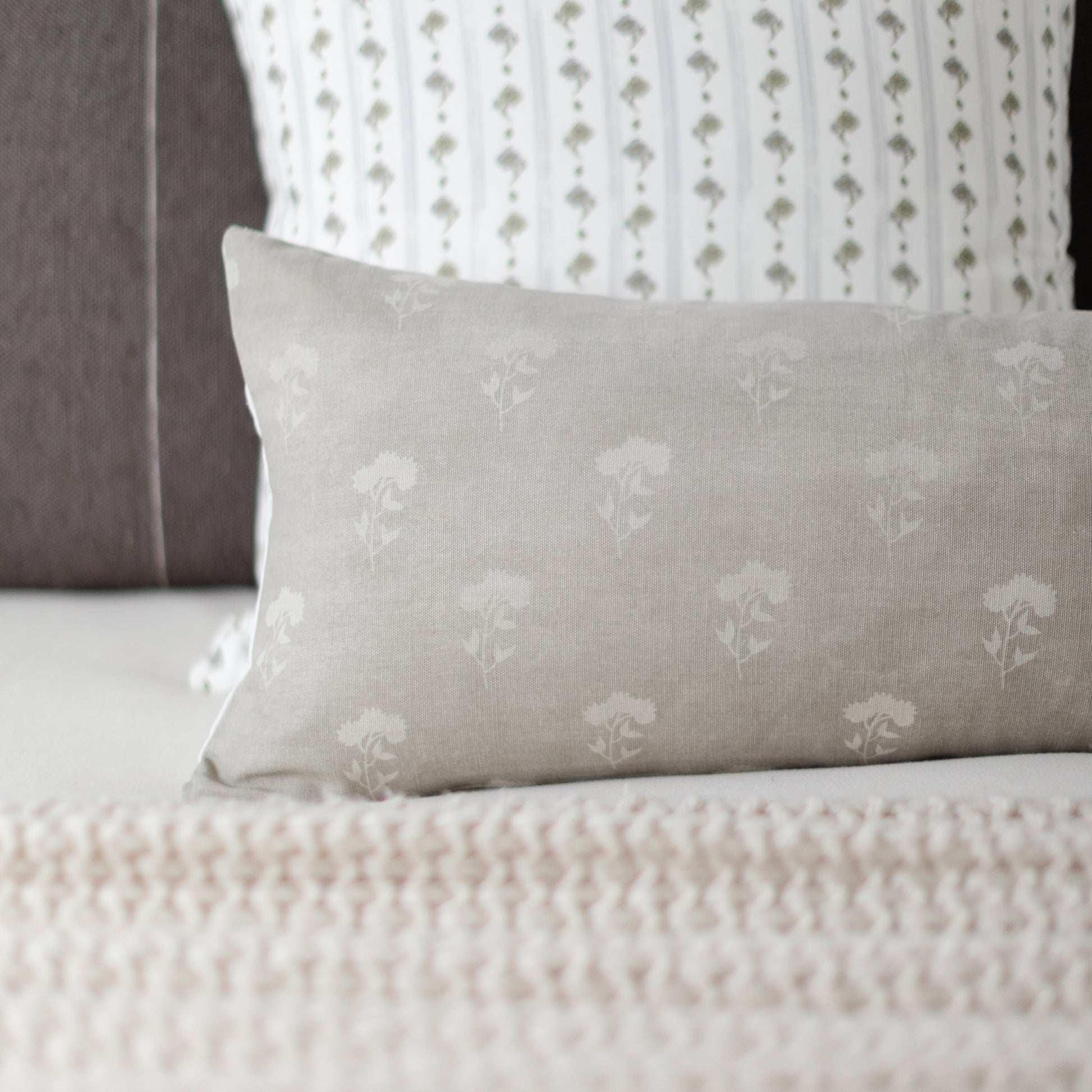 Gray lumbar pillow with white floral pattern placed on a bed with coordinating patterned pillows.