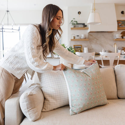 Woman arranging four decorative pillows in varying sizes on a beige sofa in a bright living room.