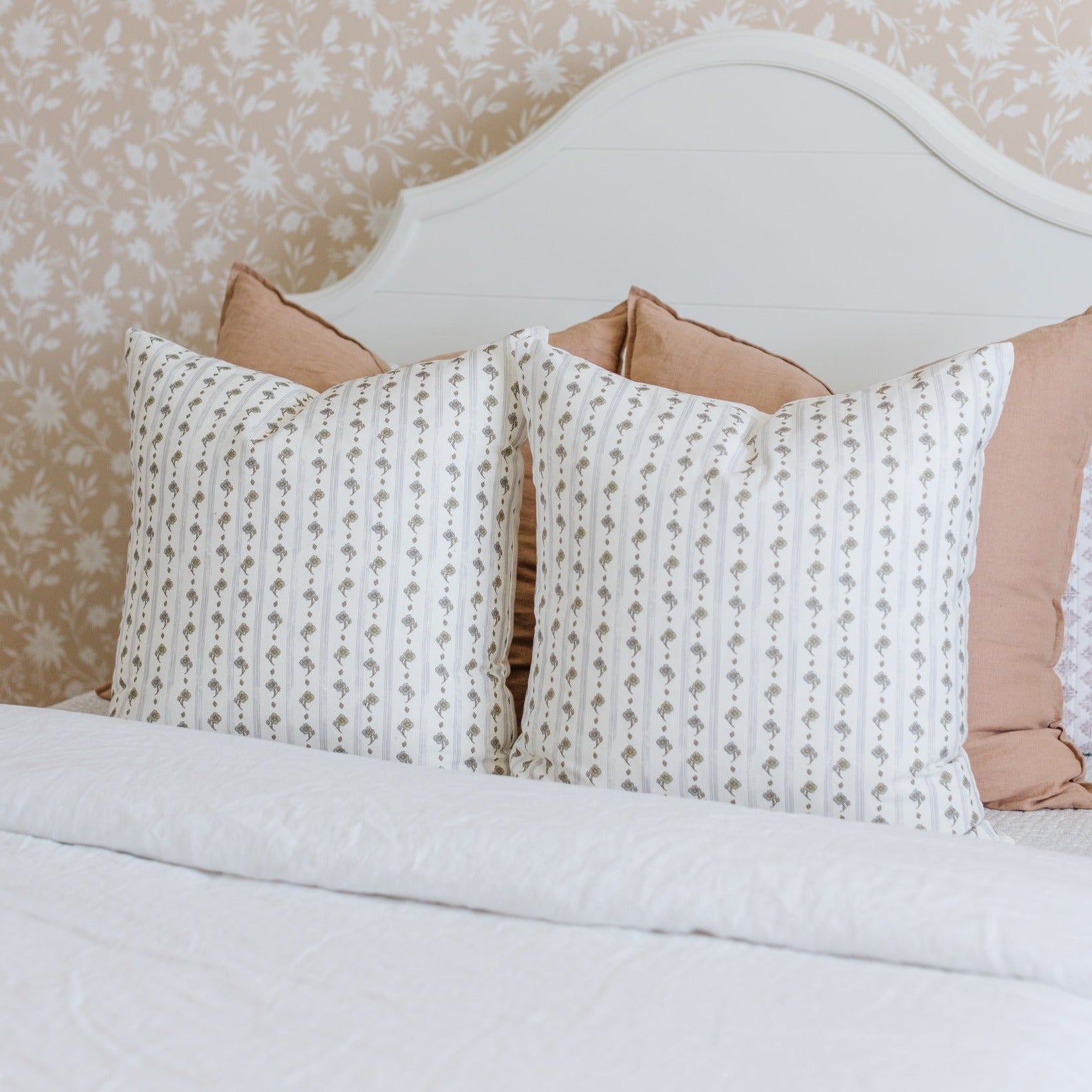 Two cream pillows with gray floral stripe pattern and tan back, displayed on white bed with curved headboard.