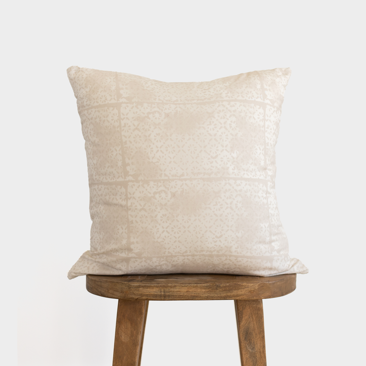 Cream-colored square pillow with faded floral pattern, displayed on wooden stool.