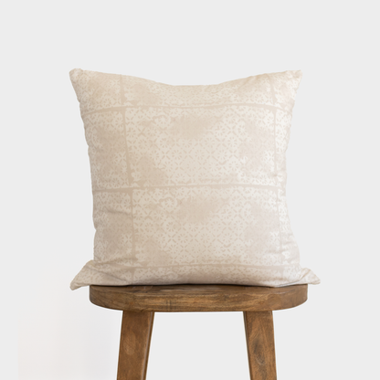 Cream-colored square pillow with faded floral pattern, displayed on wooden stool.