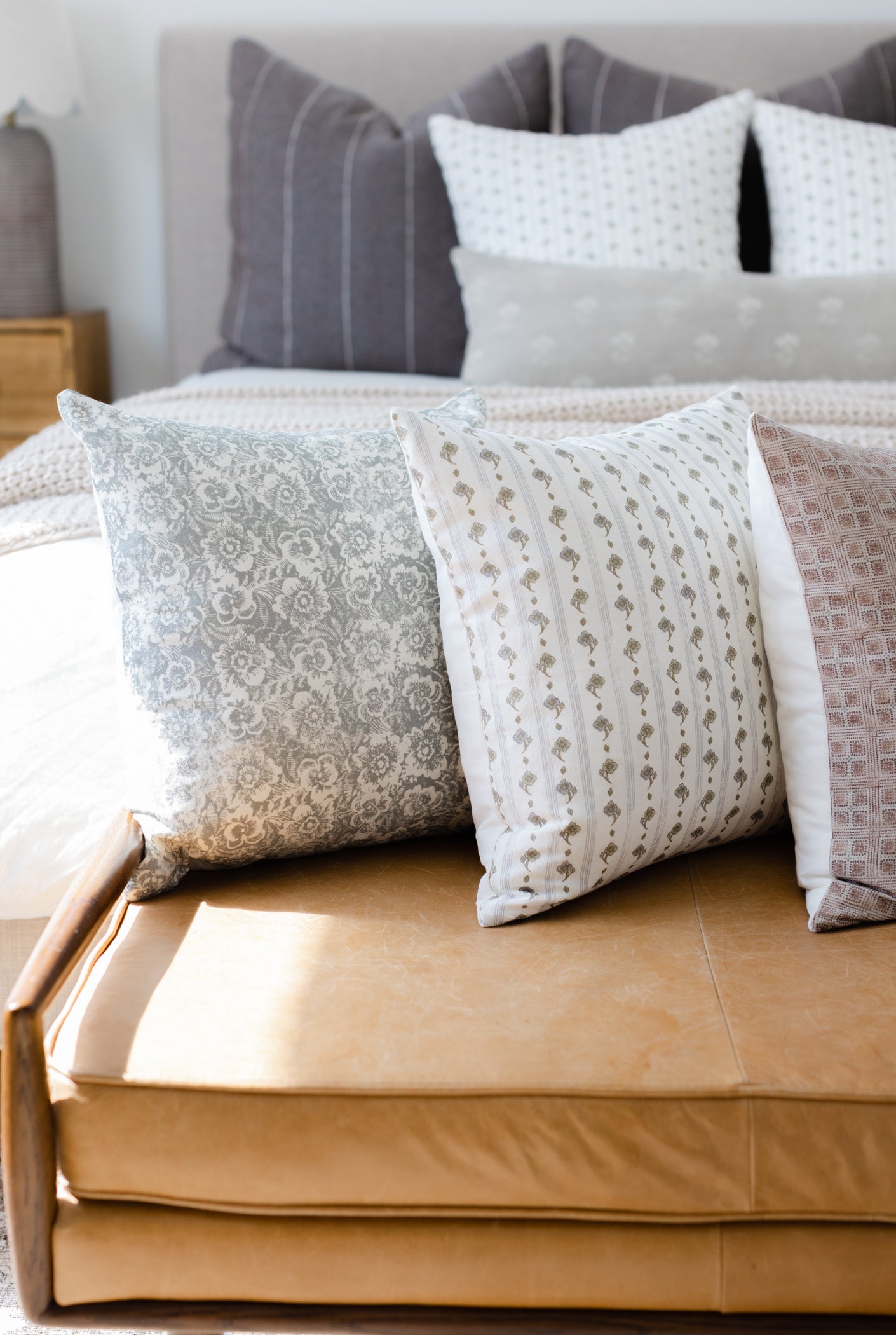 Four decorative pillows in varying patterns displayed on a tan upholstered bench in front of a bed.