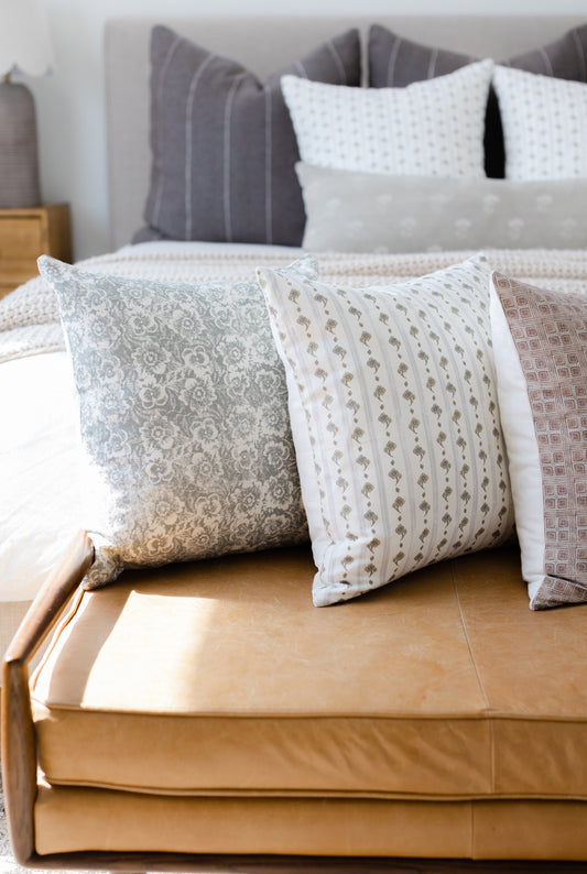 Four decorative pillows in varying patterns displayed on a tan upholstered bench in front of a bed.