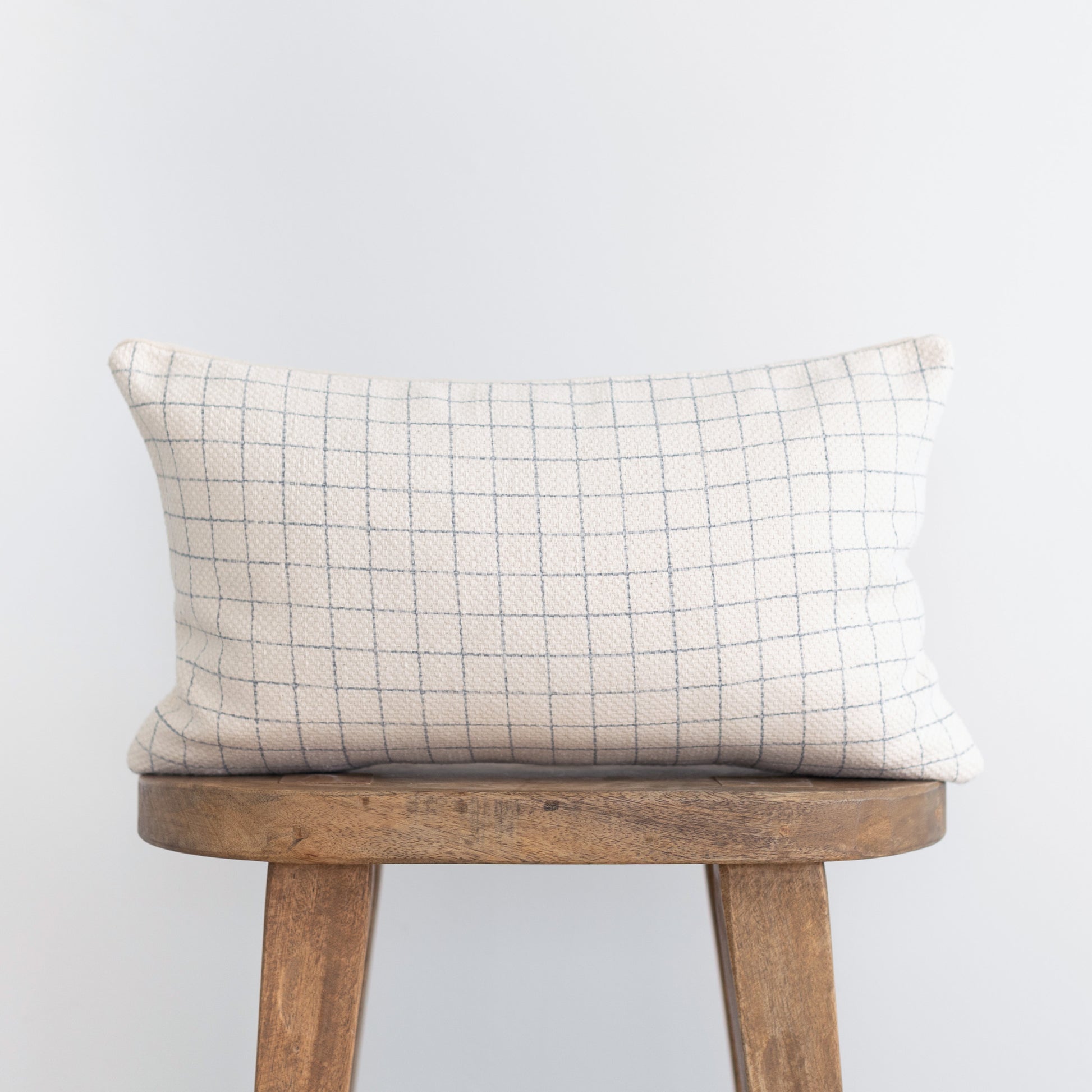 Cream lumbar pillow with gray grid pattern on wooden stool.