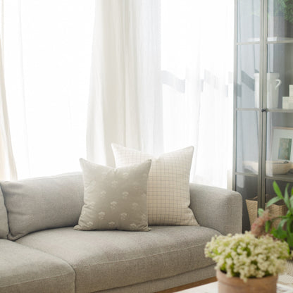 Cream-colored square pillow with grid pattern displayed on gray sofa near window.