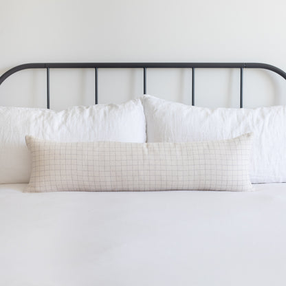 Beige lumbar pillow with grid pattern displayed on bed with white pillows and black metal headboard.