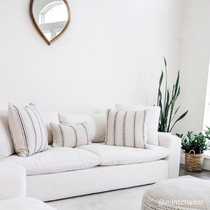 Cream linen sofa with striped throw pillows in a bright room with round mirror and potted plants.