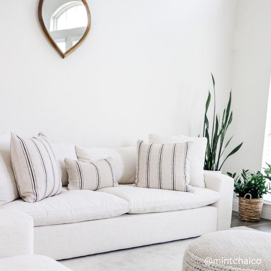 Cream linen sofa with striped throw pillows in a bright room with round mirror and potted plants.