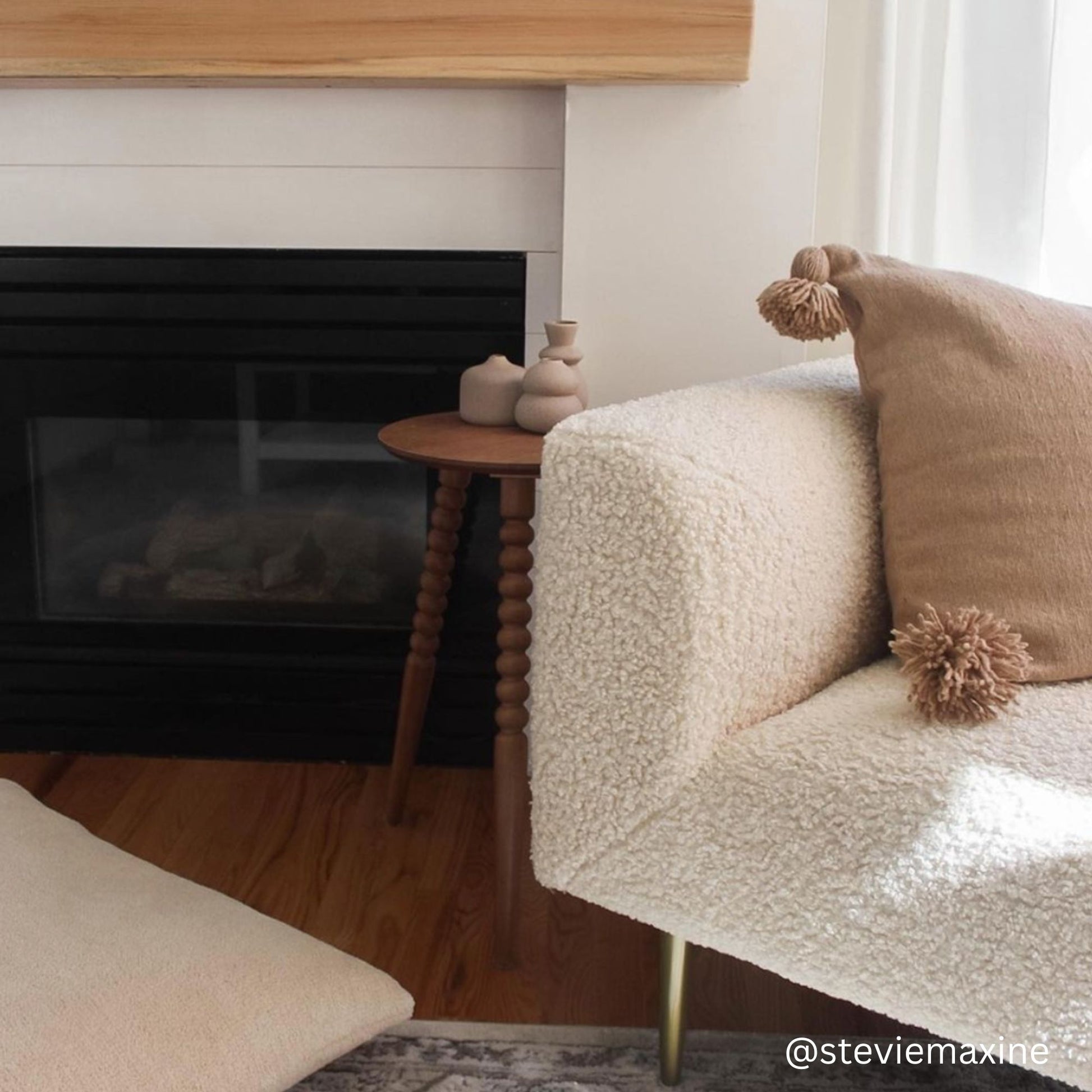Cream boucle armchair with tan pompom pillows in a living room setting next to a fireplace.